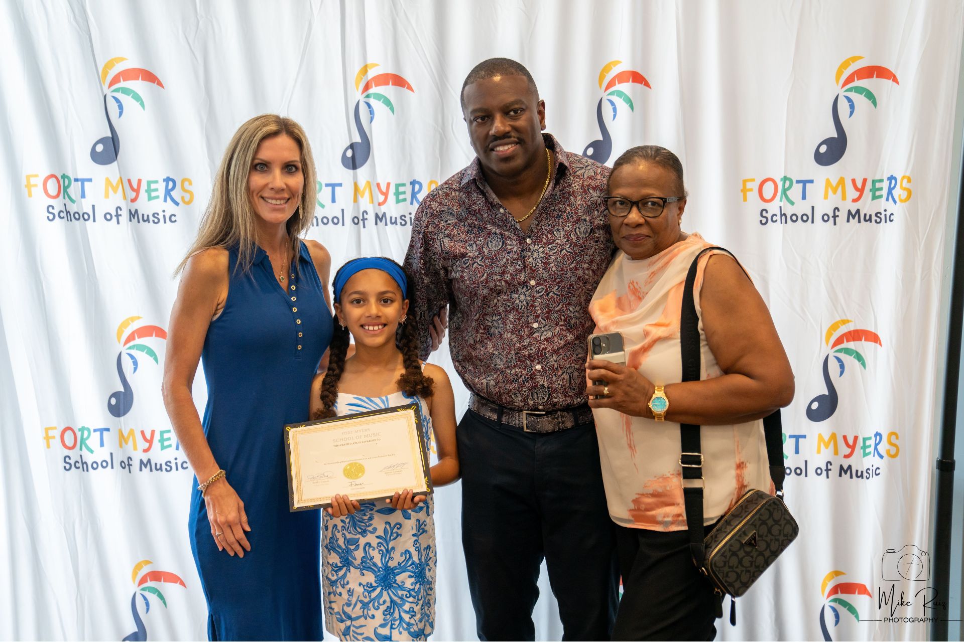 a student holding her certificate and standing with her parents and a grandparent in front of our step and repeat