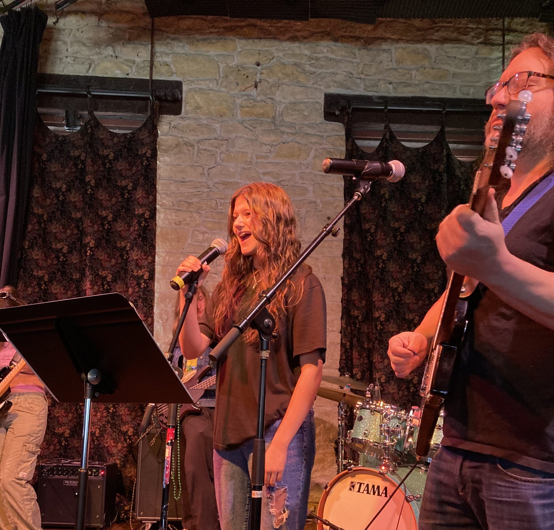 girl on stage singing into a microphone next to her guitar playing teacher